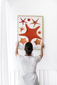 a woman is holding up a painting on the wall above her head and reaching for it