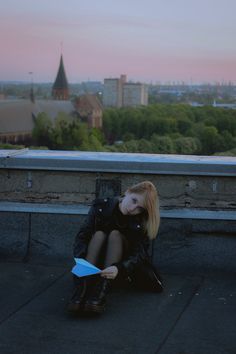 a woman sitting on the ground with a book in her hand and buildings in the background