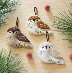 three ornaments made to look like owls hanging from christmas tree branches on a wooden table