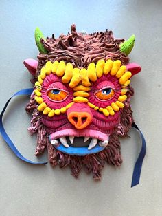 a mask made out of clay with horns and fangs on it's face, sitting next to a blue ribbon