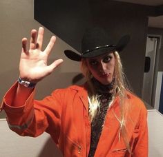 a woman in an orange jacket and hat making the vulcan sign with her hand while standing next to a wall