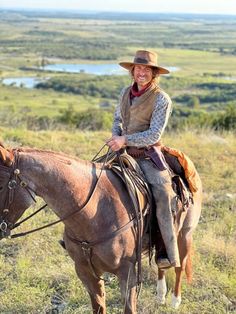 a woman riding on the back of a brown horse in a field next to a lake