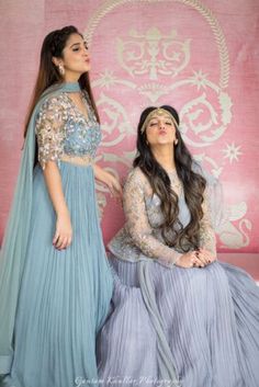 two women in long dresses sitting on a pink chair and one is wearing a blue dress