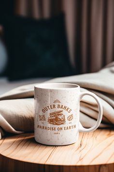 a white coffee mug sitting on top of a wooden table next to a pillow and blanket