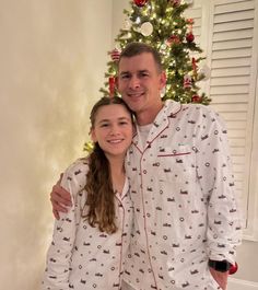 a man and woman wearing pajamas standing in front of a christmas tree with their arms around each other