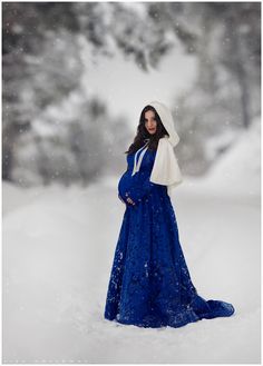 a pregnant woman standing in the snow wearing a blue dress and holding an umbrella over her head