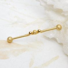 "Industrial barbell with gold stardust ball ends, bar is accented with smooth gold-filled beads, matching gold stardust center focal bead and finished off with 12k gold filled wire wrap. The center beads and wire fits snug on the bar but can be slid either direction on the bar for easy insertion. Barbell can be worn on either ear. Both of the ends on the barbell unscrew. Barbell is 14g gold steel with a 1 1/2\" wearable bar length.  *Wearable bar length is measured between the ball ends! For Ind Industrial Barbell, Industrial Piercing, Tanning Lotion, Body Jewellery, Stardust, Focal Bead, Beads And Wire, Piercing Jewelry, Jewelry Sales