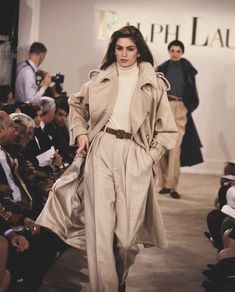 a woman is walking down the runway in a trench coat and pants, while others watch