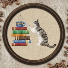 a cross stitch picture with coffee and a cat drinking from a cup on top of books