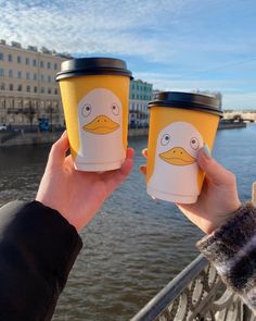 two people holding up coffee cups in front of the water with ducks painted on them