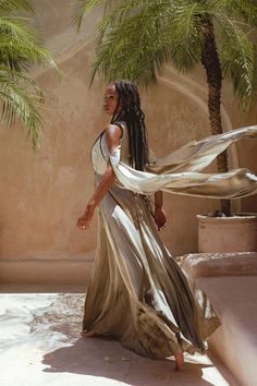 a woman with dreadlocks is standing in front of a palm tree wearing a flowing dress