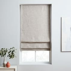 a white room with a window covered in roman shades and a vase filled with flowers