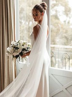 a woman in a wedding dress is standing by a window with her veil over her head