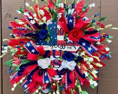 a patriotic wreath with flowers and ribbons hanging on a wooden wall, decorated with the american flag