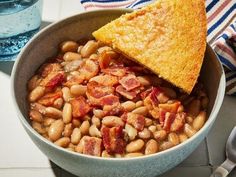 a bowl filled with beans and bacon next to a piece of bread on top of a table