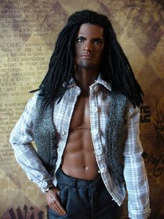 a male doll with long black hair wearing a shirt and pants, standing in front of a wall