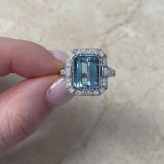 This stunning ring showcases a captivating 3.27 carats of emerald-cut aquamarine at its center. elegantly bezel-set and enhanced by a halo of old European cut diamonds; flanking the center stone are baguette-cut diamonds, adding to the overall allure. Each shoulder of the ring is adorned with a row of old Euro[pean cut diamonds. The total diamond weight of the ring is approximately 0.62 carats accentuating its brilliance.  The ring is handcrafted in platinum and features delicate milgrain detail Heirloom Diamond Topaz Ring With Emerald Cut, Heirloom Diamond Topaz Ring, Emerald Cut, Emerald-cut Topaz Ring With Halo Setting, Baguette Cut Topaz Ring With Diamond Halo Setting, Baguette Cut Topaz Ring With Diamond Halo, Emerald Cut Topaz Ring In Platinum, Platinum Topaz Ring Emerald Cut, Gia Certified Emerald Cut Aquamarine Ring, Gia Certified Emerald Cut Topaz Ring In Platinum