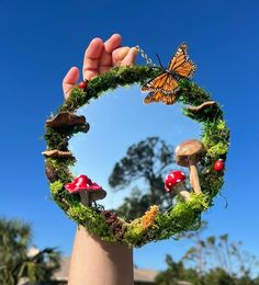 a hand holding up a mirror with mushrooms and butterflies on it, in front of a blue sky