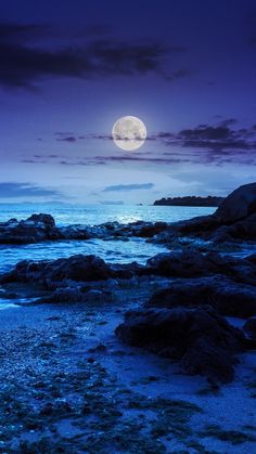 the full moon is setting over the water and rocks on the beach at night time