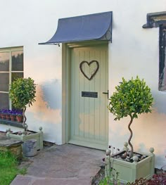 two potted plants in front of a white house with a heart shaped sign on the door