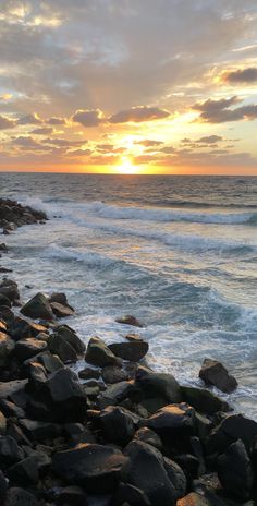 the sun is setting over the ocean with rocks on the shore and waves coming in