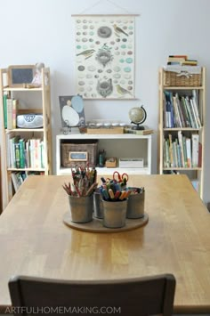 a wooden table topped with two buckets filled with markers and pencils next to bookshelves