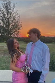 a man and woman standing next to each other in front of a field at sunset