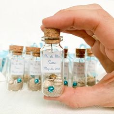 a hand holding a tiny glass bottle filled with sand and sea shells in front of small bottles