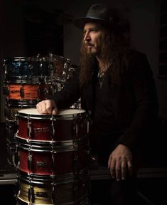 a man with long hair sitting next to a drum set in front of a dark background