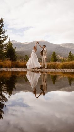 a bride and groom dancing in front of a lake with mountains in the back ground