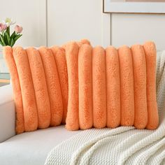 four orange pillows on a white couch with flowers in the corner and a pink flower pot behind them