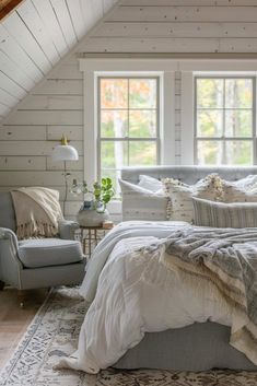 a bed sitting under two windows in a bedroom next to a chair and ottoman with pillows on it