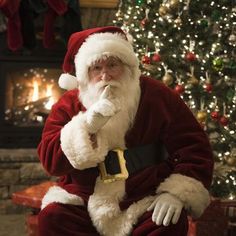 a man dressed as santa claus sitting in front of a christmas tree with his hand on his chin