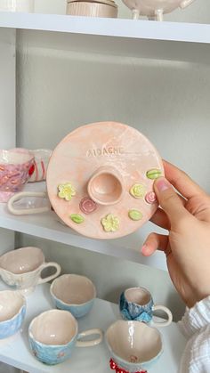 a person is holding a ceramic object in front of some cups and saucers on a shelf