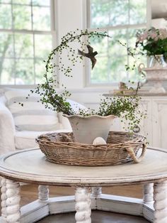 a white table topped with a basket filled with flowers and greenery next to a window