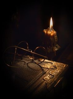 a lit candle sitting on top of an old book with glasses around it in the dark