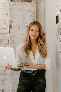 a woman standing in front of a white brick wall holding a laptop computer and looking at the camera