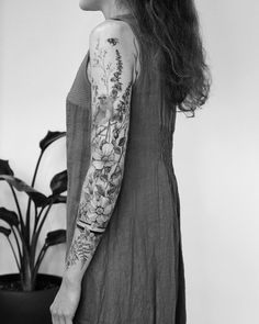 a woman with long hair and tattoos on her arm standing in front of a potted plant