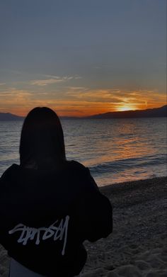 a person is standing on the beach watching the sun go down over the water and mountains