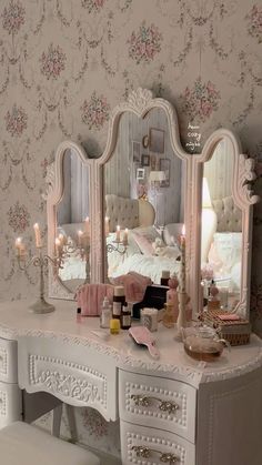 a white dresser with two mirrors and candles on it in front of a floral wallpapered bedroom