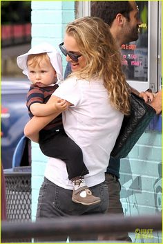 a woman holding a small child in her arms while standing next to a man with sunglasses on