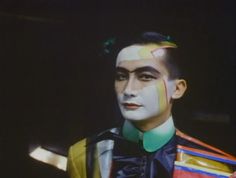 a man with multicolored hair and makeup looks at the camera while wearing a jacket