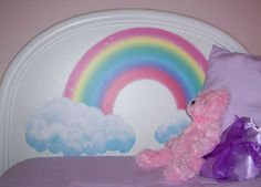 a pink teddy bear sitting on top of a bed next to a rainbow wall decal