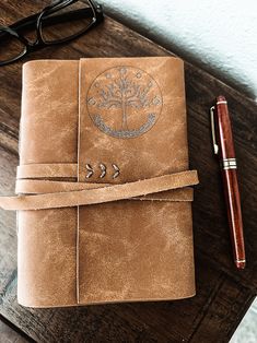 a brown leather journal sitting on top of a wooden table next to a pen and glasses