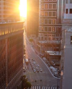 the sun is setting over a city street with tall buildings and cars driving down it