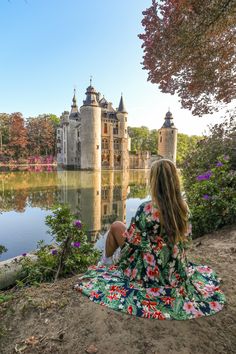 a woman sitting on the ground in front of a castle with water and flowers around her