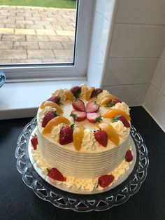 a white cake with fruit on top sitting in front of a window sill next to a glass pane