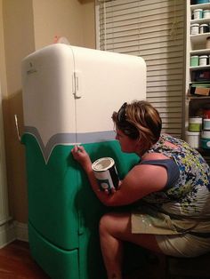 a woman is painting the side of a refrigerator with green paint and she is holding a can