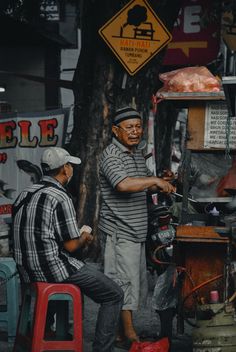 two men are sitting on stools in front of a food stand and one man is pointing at something