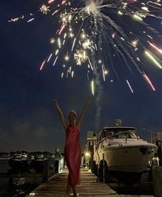 a woman in a red dress is standing on a dock with her arms up as fireworks go off behind her